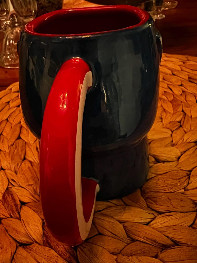 Puede incluir: Una taza de cer&aacute;mica con exterior azul marino e interior rojo. El asa es roja con una franja blanca. La taza se encuentra sobre una superficie tejida y texturizada. La taza es una pieza &uacute;nica para beber.