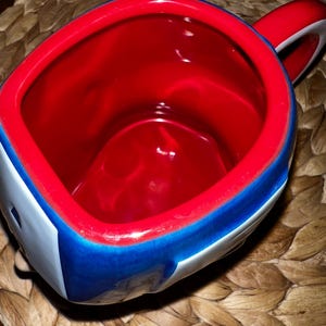 Puede incluir: Una taza de cer&aacute;mica con interior rojo y borde rojo. El exterior presenta un dise&ntilde;o azul y blanco. El asa es roja y blanca. La taza est&aacute; sobre una superficie tejida.