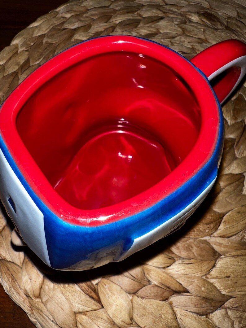 Puede incluir: Una taza de cer&aacute;mica con interior rojo y borde rojo. El exterior presenta un dise&ntilde;o azul y blanco. El asa es roja y blanca. La taza est&aacute; sobre una superficie tejida.