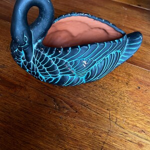 May include: A black swan-shaped planter with turquoise accents. The ceramic planter has a textured surface and a terracotta interior. The swan's neck forms a handle, and the wings are detailed with carved feathers. A decorative piece for plants.