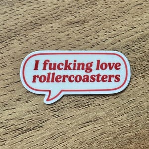 May include: A white speech bubble sticker with a red border and red text that reads "I fucking love rollercoasters" in a bold, sans-serif font. The sticker has a rounded, elongated shape with a small tail pointing downward on the left side.