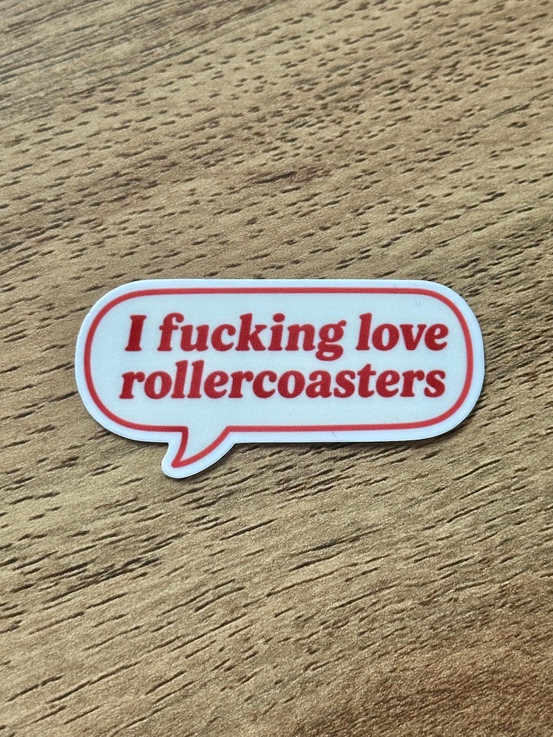 May include: A white speech bubble sticker with a red border and red text that reads "I fucking love rollercoasters" in a bold, sans-serif font. The sticker has a rounded, elongated shape with a small tail pointing downward on the left side.