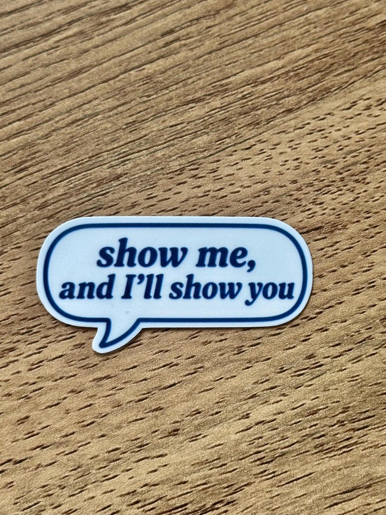 May include: A white speech bubble sticker with a dark blue border and the dark blue text "show me, and I'll show you" printed in the center. The sticker has a rounded rectangular shape with a small triangular tail pointing downward.