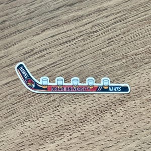 May include: A die-cut sticker shaped like a hockey stick with a red, white, and blue color scheme. The design features a hawk mascot head, the text HAWKS, and the text BRIAR UNIVERSITY. Five small clear shot glasses are lined up along the blade.