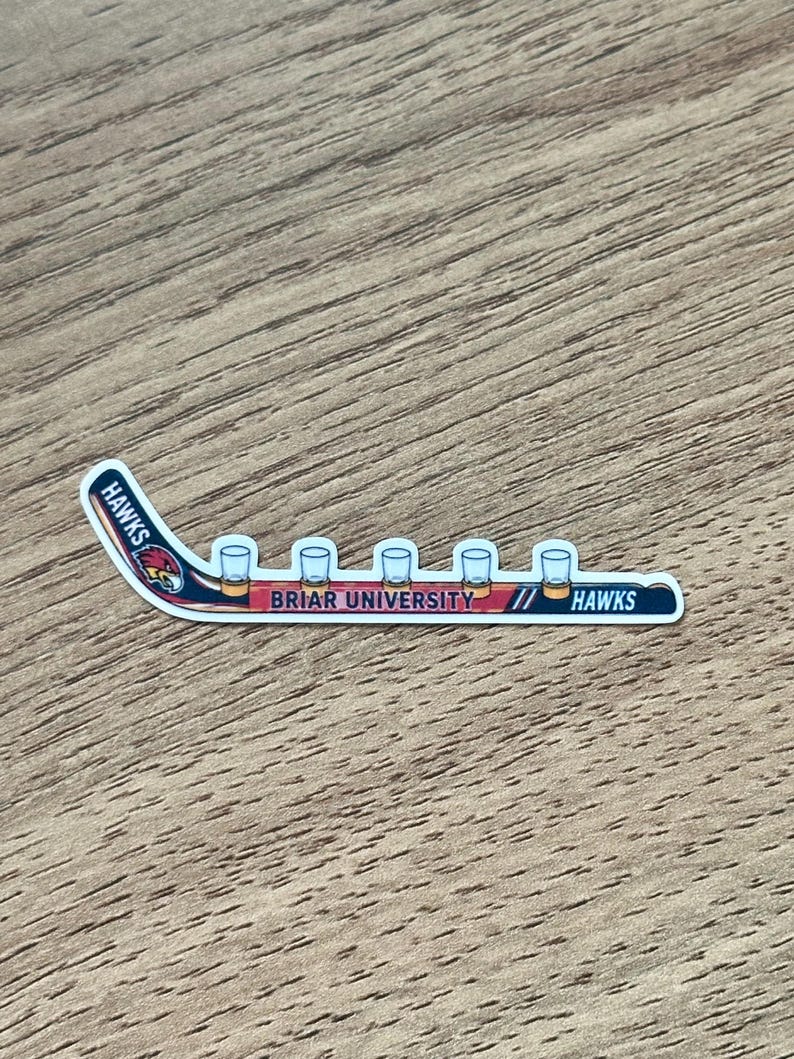 May include: A die-cut sticker shaped like a hockey stick with a red, white, and blue color scheme. The design features a hawk mascot head, the text HAWKS, and the text BRIAR UNIVERSITY. Five small clear shot glasses are lined up along the blade.