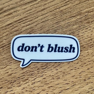 May include: A white speech bubble sticker with a dark blue outline and the text "don't blush" printed in dark blue lowercase letters. The sticker has a rounded rectangular shape with a small tail pointing downwards on the left side.