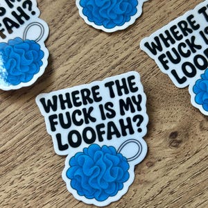 Peut inclure: Plusieurs autocollants blancs avec l'inscription "WHERE THE FUCK IS MY LOOFAH ?" et un motif de loofah bleu. Les autocollants sont posés sur une surface en bois. Les autocollants sont découpés.