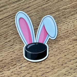 May include: A sticker featuring a black hockey puck with two rabbit ears attached to the top. The ears are light blue on the outer edges with bright pink inner sections. One ear stands upright while the other ear is folded over to the side.