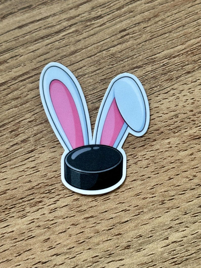 May include: A sticker featuring a black hockey puck with two rabbit ears attached to the top. The ears are light blue on the outer edges with bright pink inner sections. One ear stands upright while the other ear is folded over to the side.