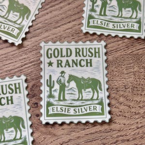 Puede incluir: Varias pegatinas verdes y blancas con un diseño de sello postal. Cada sello presenta un vaquero con un caballo, un cactus y las palabras "Gold Rush Ranch" y "Elsie Silver".