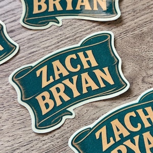 Op de afbeelding: Verschillende stickers met de naam "ZACH BRYAN" in gouden letters op een teal banner-ontwerp. De stickers hebben een vintage uitstraling, met een crème rand. Ze liggen verspreid op een lichtbruin houten oppervlak.