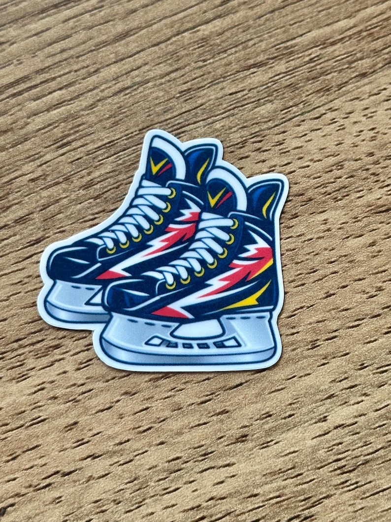 May include: A die-cut sticker featuring a pair of dark blue ice skates with white laces and a lightning bolt graphic in red and yellow on the side. The skates have silver blades and are outlined in white, presented as a single graphic illustration.