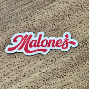 May include: A die-cut sticker featuring the word Malone's written in a bold, red, cursive script font with a white outline. The lettering has a smooth, rounded appearance and is presented against a plain, light-colored background surface.
