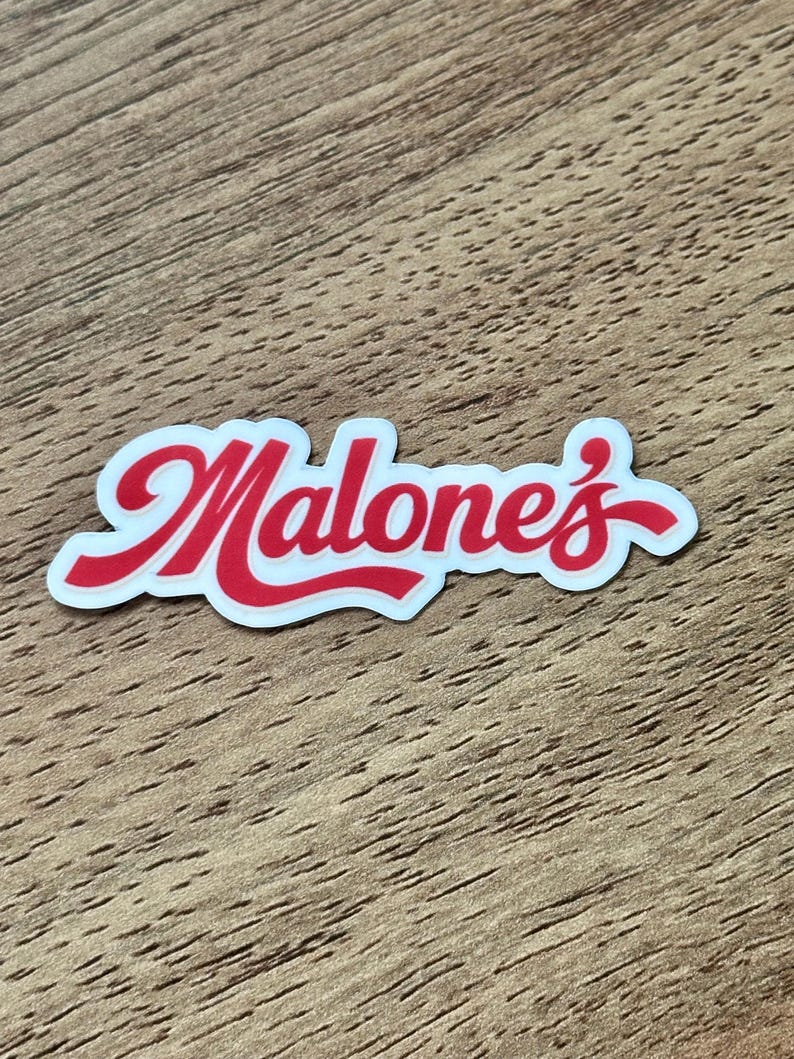 May include: A die-cut sticker featuring the word Malone's written in a bold, red, cursive script font with a white outline. The lettering has a smooth, rounded appearance and is presented against a plain, light-colored background surface.
