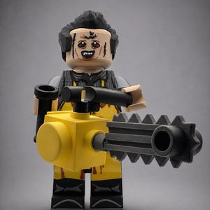 May include: A plastic minifigure featuring a mask-like face with dark hair, wearing a grey shirt under a yellow apron with red blood-like splatters, black pants, and holding a large yellow and black toy chainsaw with a serrated blade.