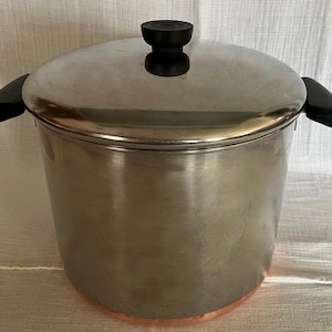 Puede incluir: Una olla de acero inoxidable con tapa a juego, que cuenta con dos asas laterales negras y un pomo negro en la tapa. La olla tiene un cuerpo cilíndrico con un acabado metálico pulido y una base de color cobre visible en el borde inferior.