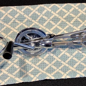 May include: A vintage chrome hand mixer with black handles. The mixer features a gear mechanism and a rotating beater design. The mixer is lying on a light blue and white checkered patterned cloth.