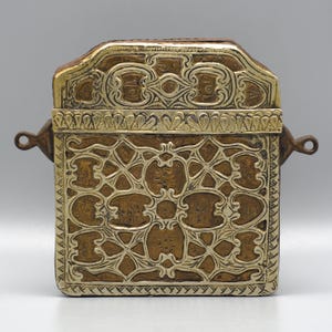 Puede incluir: Una caja rectangular antigua con intrincados diseños de filigrana dorada y plateada. La caja tiene una tapa con bisagras y dos pequeñas asas en los laterales. La orfebrería presenta patrones florales y geométricos.
