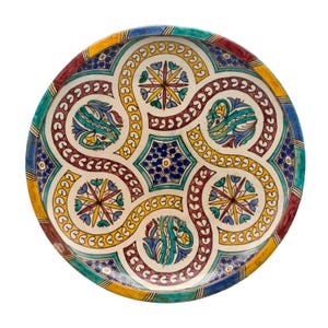 May include: A round, decorative ceramic plate with a complex, colourful design. The plate features a white background with a repeating pattern of yellow, blue, red, and teal shapes. The rim has a striped pattern in various colours. The plate is likely handmade.