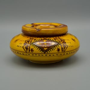 May include: A yellow ceramic ashtray with a lid. The ashtray has a round shape with a decorative pattern in brown and dark yellow. The lid has a circular design with a groove for resting cigarettes.
