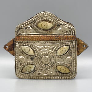 Puede incluir: Una caja rectangular plateada con incrustaciones decorativas en forma de ojo. La caja presenta una tapa con bisagras y una banda de color cobre. Los bordes están adornados con pequeños adornos circulares. La caja tiene una estética vintage o antigua.