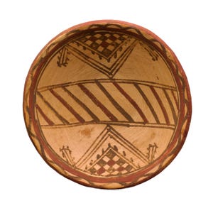 May include: A round, beige ceramic bowl with a red rim and geometric patterns. The interior features alternating red and brown diagonal stripes and triangular designs with checkered squares. The bowl's design is symmetrical and centered.