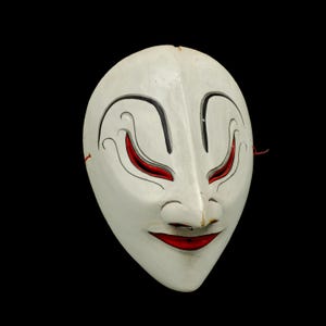 May include: A white wooden mask with black and red painted details. The mask has a wide, open mouth with a red smile and large, almond-shaped eyes with red pupils. The mask is a traditional Japanese Noh mask.