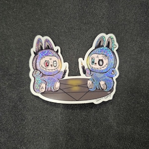 May include: A sticker featuring two cartoon figures in blue bunny suits with glitter accents. Each figure holds a black candle and sits at a table with a pentagram design. The sticker has a white border and a dark background.