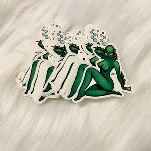 May include: A stack of stickers featuring a green creature with a fish-like head and body, with bubbles above its head. The creature is in a pin-up pose. The stickers are on a white, fluffy surface.