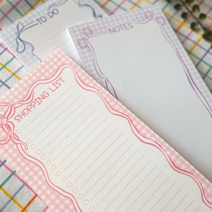 May include: Three notepads with decorative borders and text. The notepad in the foreground has a pink gingham pattern and the words "SHOPPING LIST". The other notepads have the words "TO DO" and "NOTES".