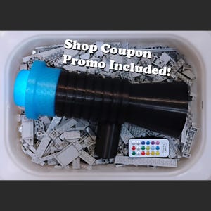 Puede incluir: Una pistola de juguete negra y azul con una boquilla azul claro y un mando a distancia con botones de colores. La pistola de juguete está rodeada de bloques de construcción grises. El texto en la imagen dice "Shop Coupon Promo Included!"