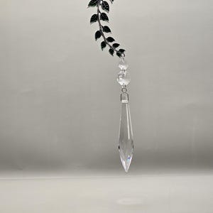 May include: A decorative hanging ornament featuring a silver-toned vine with green leaves and clear crystal accents. The ornament has a long, pointed crystal pendant at the bottom, adding a touch of elegance. The design is suitable for home decor.