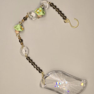 May include: A decorative suncatcher with a clear, leaf-shaped crystal pendant. The suncatcher features a chain of small, brown beads, clear crystal beads, and green mushroom-shaped charms. A gold-colored hook is at the top for hanging.