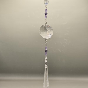 May include: A clear crystal suncatcher with a large, round, faceted crystal disc and a teardrop-shaped pendant. The suncatcher is strung with purple beads and smaller clear crystals, creating a decorative hanging ornament.