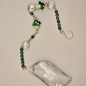 May include: A decorative suncatcher with a clear, leaf-shaped crystal and a gold-colored hook. The suncatcher features green beads, small mushroom charms with white polka dots, and clear crystal beads.
