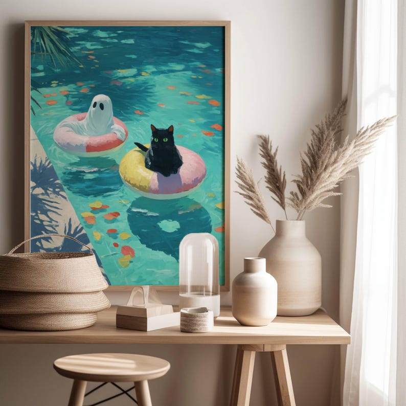 Op de afbeelding: Een ingelijste kunstafdruk toont een zwarte kat en een spook die in een turquoise zwembad op opblaasbare ringen drijven. Er liggen kleurrijke bladeren op het water. Het kunstwerk wordt getoond op een houten tafel met decoratieve items.
