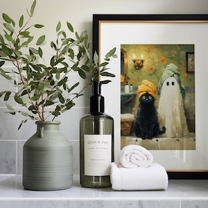 May include: A bathroom scene featuring a framed art print of a black cat and ghost wearing towels, a green vase with leafy branches, a soap dispenser, and a rolled white towel. The art print is in a black frame.