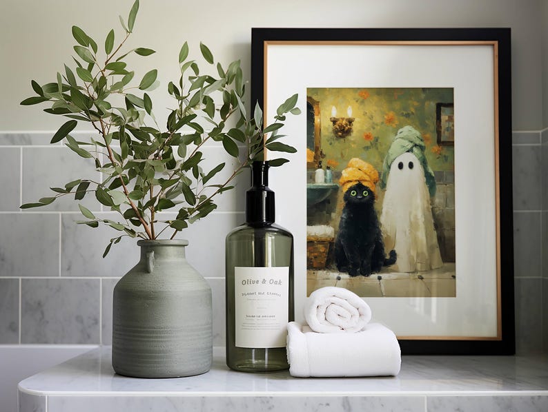 May include: A bathroom scene featuring a framed art print of a black cat and ghost wearing towels, a green vase with leafy branches, a soap dispenser, and a rolled white towel. The art print is in a black frame.