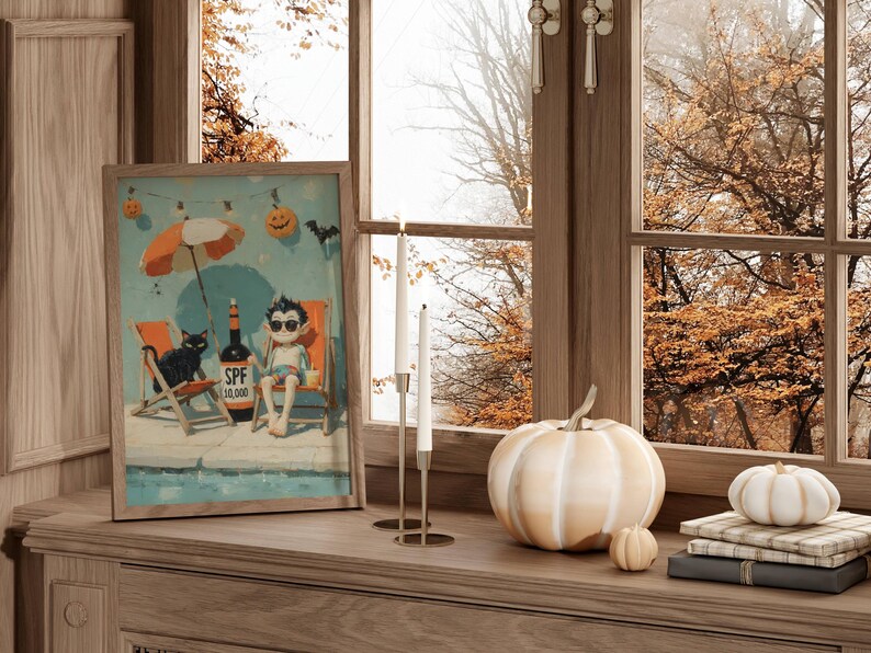 May include: A framed print depicts a cartoon scene of a vampire relaxing at a pool with a black cat, a bottle of SPF 10,000, and Halloween decorations. A white pumpkin, two smaller pumpkins, and books sit on a wooden surface near a window.