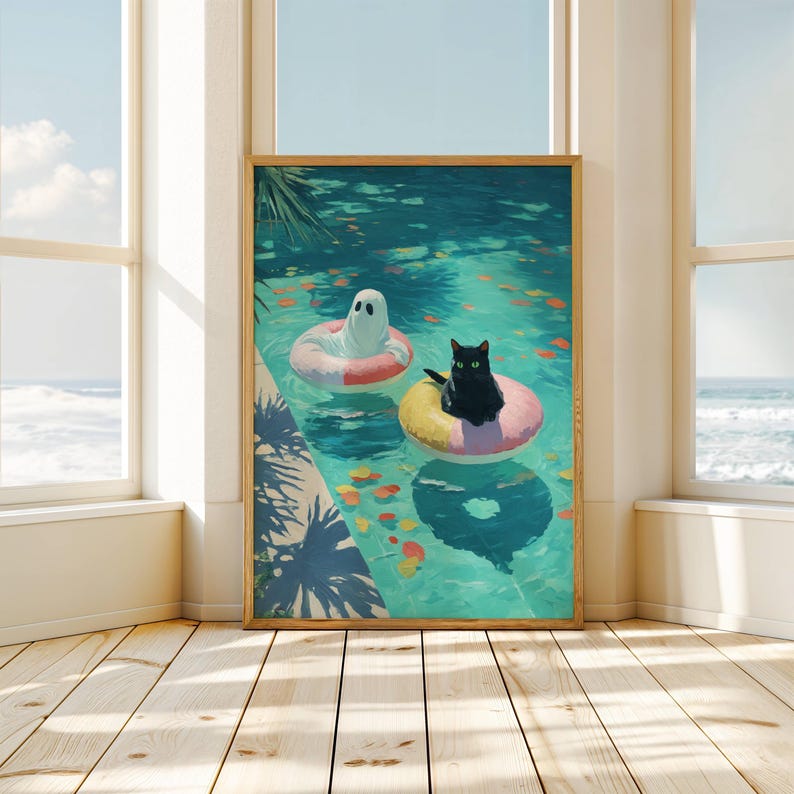 Op de afbeelding: Een ingelijste kunstdruk toont een zwarte kat en een geest die in een turquoise zwembad op opblaasbare ringen drijven. Er drijven kleurrijke bladeren in het water. Het kunstwerk wordt in een lichte kamer getoond.