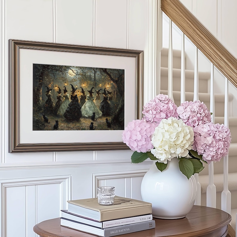 May include: Framed artwork depicting a gathering of witches in a moonlit forest, with black cats. The artwork is in a gold frame, mounted on a white paneled wall. A white vase holds pink and white hydrangeas, and books are on a wooden table.