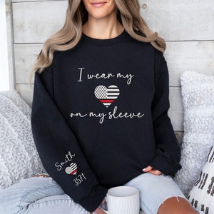 May include: Black sweatshirt with the text "I wear my heart on my sleeve" and a red line heart with an American flag design. The left sleeve has the text "Smith 3571" and a smaller red line heart with an American flag design.