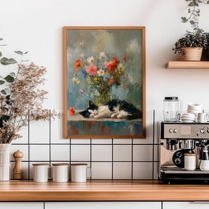 May include: A framed painting depicts a black and white cat resting near a vase of colorful flowers. The artwork hangs above a wooden countertop with a coffee machine, mugs, and other kitchen items. The background is a white tiled wall.