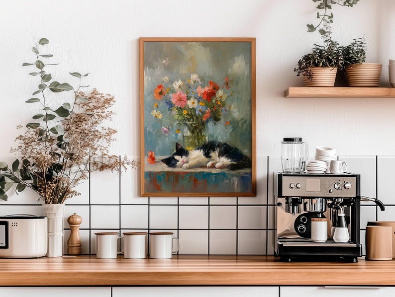 May include: A framed painting depicts a black and white cat resting near a vase of colorful flowers. The artwork hangs above a wooden countertop with a coffee machine, mugs, and other kitchen items. The background is a white tiled wall.