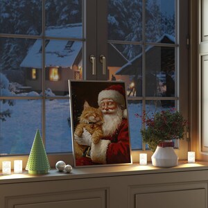 May include: A framed painting of Santa Claus holding a ginger cat. The painting is on a windowsill with a small green Christmas tree and white candles.