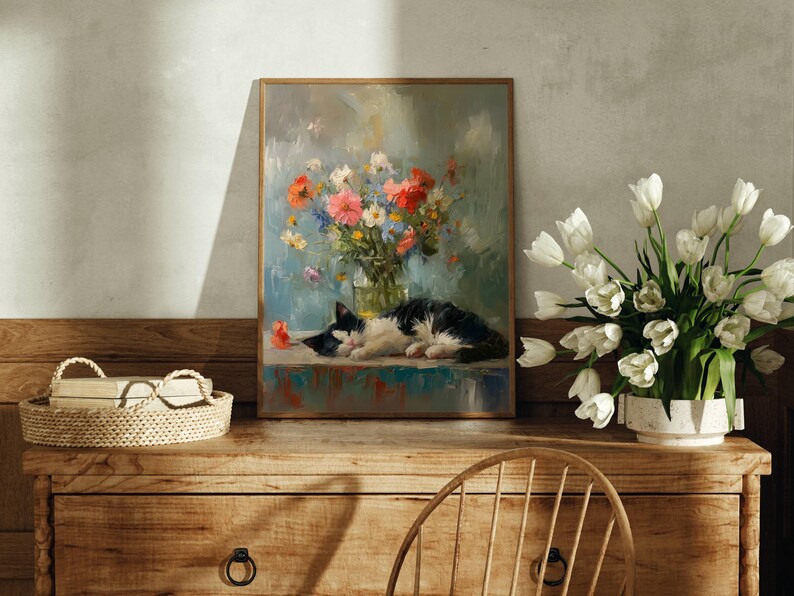 May include: A framed painting of a black and white cat sleeping next to a vase of colorful flowers. A bouquet of white tulips sits in a white vase on a wooden dresser. A woven basket with glasses rests on the dresser.
