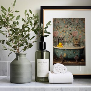 May include: A framed print of a cat in a bathtub with a rubber ducky. The print is on a white background with a floral pattern. The frame is black with a gold trim. A green vase with a leafy branch and a clear glass bottle with a black label are on a white countertop.