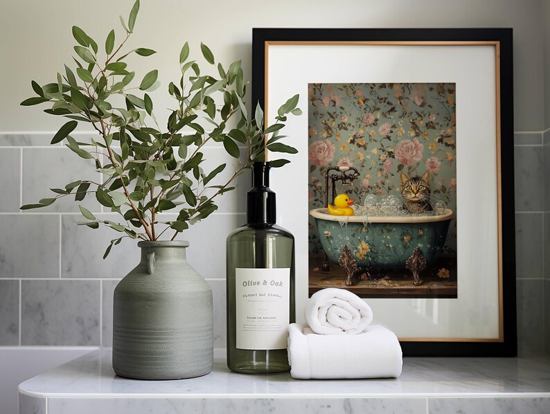May include: A framed print of a cat in a bathtub with a rubber ducky. The print is on a white background with a floral pattern. The frame is black with a gold trim. A green vase with a leafy branch and a clear glass bottle with a black label are on a white countertop.
