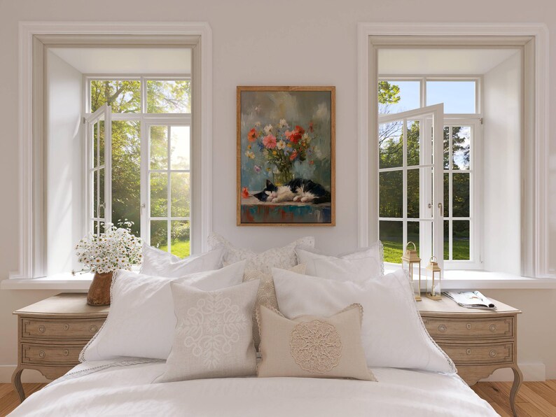 May include: A bedroom scene featuring a bed with white bedding and decorative pillows. A framed painting of a cat and flowers hangs above the bed. Two windows with open panes offer a view of a lush green landscape.