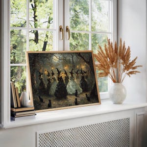 May include: A framed print of a gathering of witches in a moonlit forest. The artwork features figures in long gowns and pointed hats, with black cats scattered around. The print is displayed on a white windowsill with a vase of dried plants.